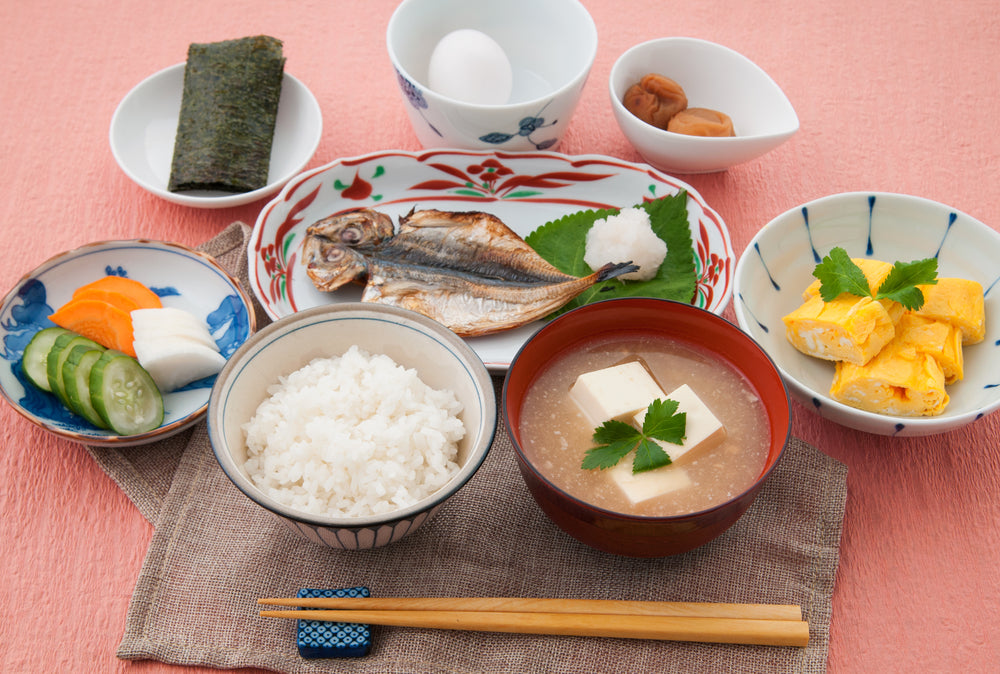 A traditional Japanese breakfast with miso soup, umeboshi plums, white rice, oshinko Japanese pickles, tamagoyaki rolled omelet, and fish.